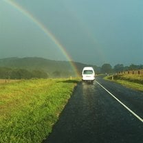 A white van is driving down a local road. There is a rainbow coming from the left to meet the van.